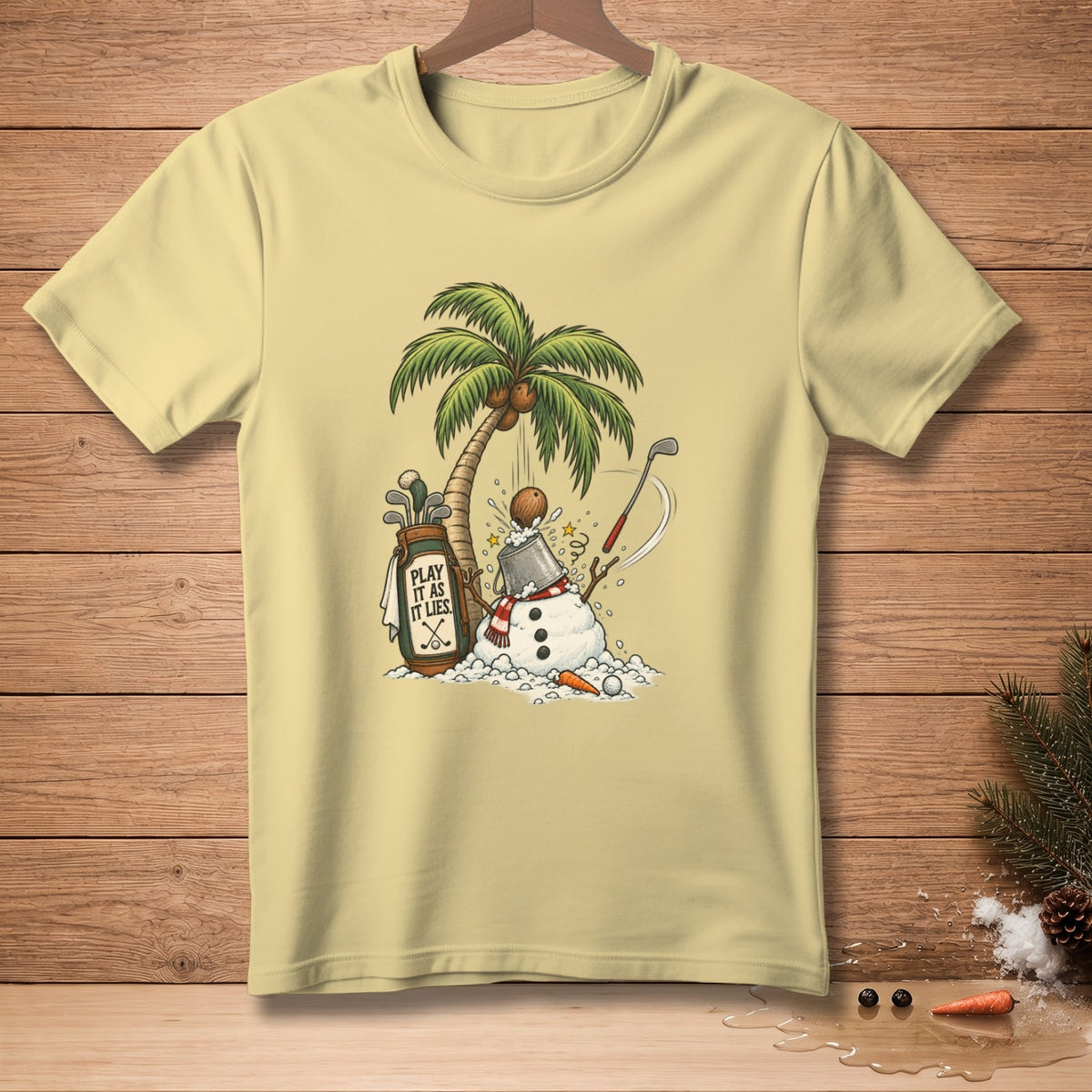 Light yellow t-shirt with snowman, palm tree, golf bag, and "Play It As It Lies" text.