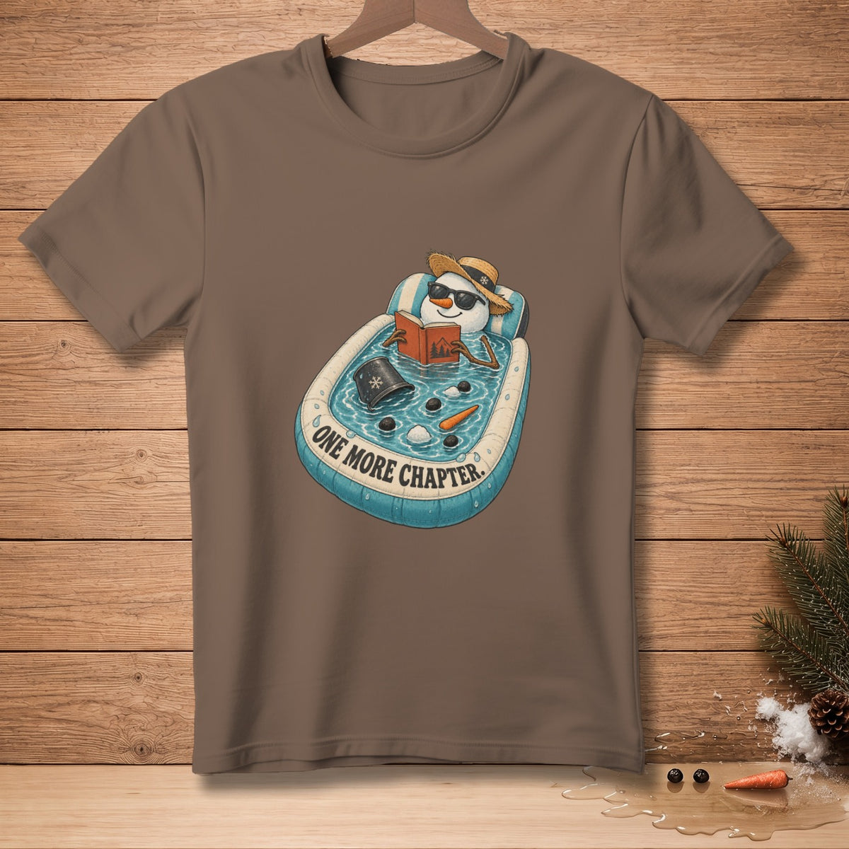 T-shirt with a snowman relaxing in a pool reading a book, text "One More Chapter".