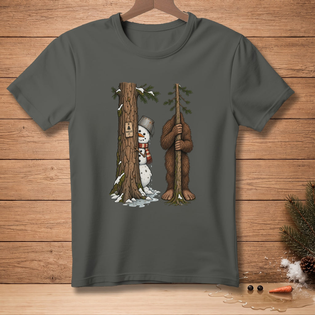 Bigfoot holds a small tree hiding a snowman peeking from behind a larger tree.