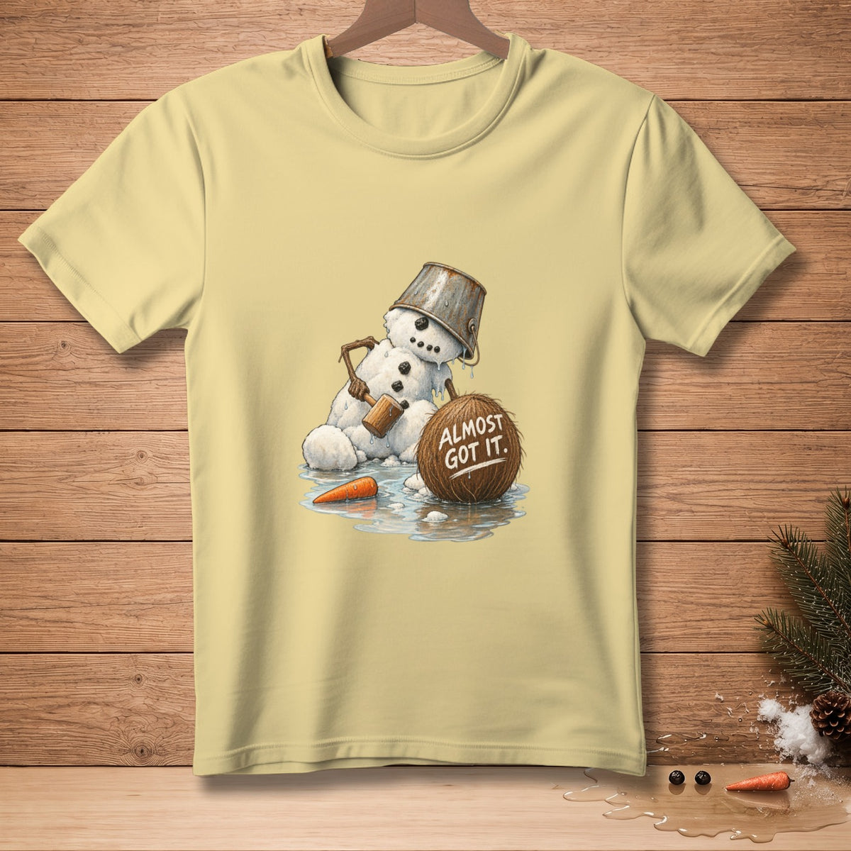 T-shirt graphic: melting snowman with bucket hat, holding hammer, next to coconut with text "ALMOST GOT IT."