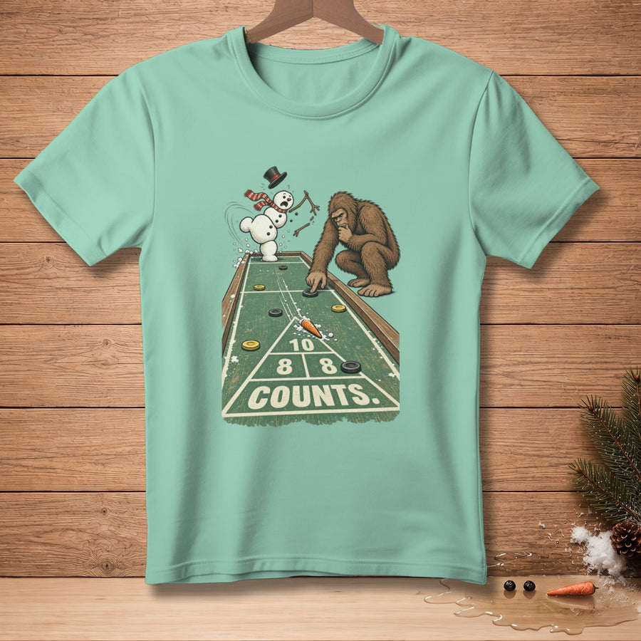 Bigfoot and snowman play shuffleboard on t-shirt graphic with "COUNTS" text.