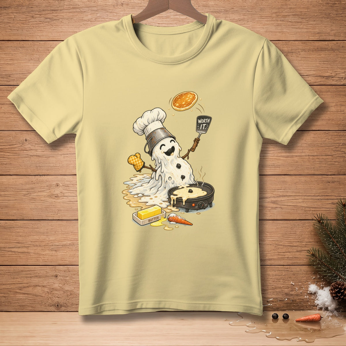 Melting snowman chef flips a pancake with "Worth It" spatula on a light yellow t-shirt.