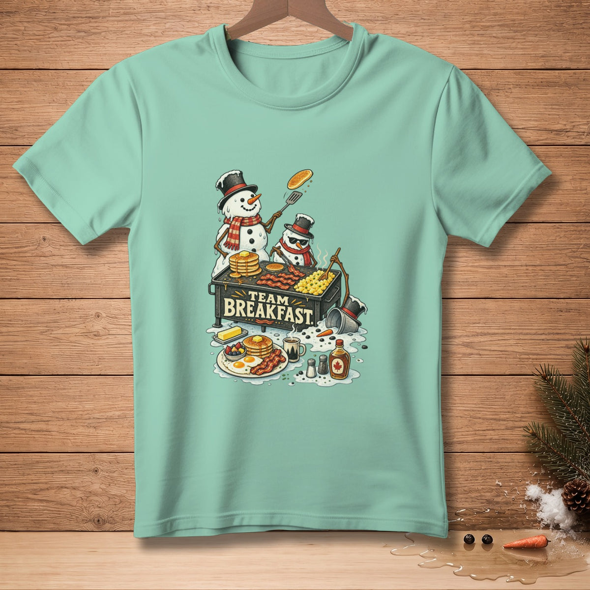Snowmen cooking breakfast on a griddle with text "TEAM BREAKFAST" on a mint t-shirt.
