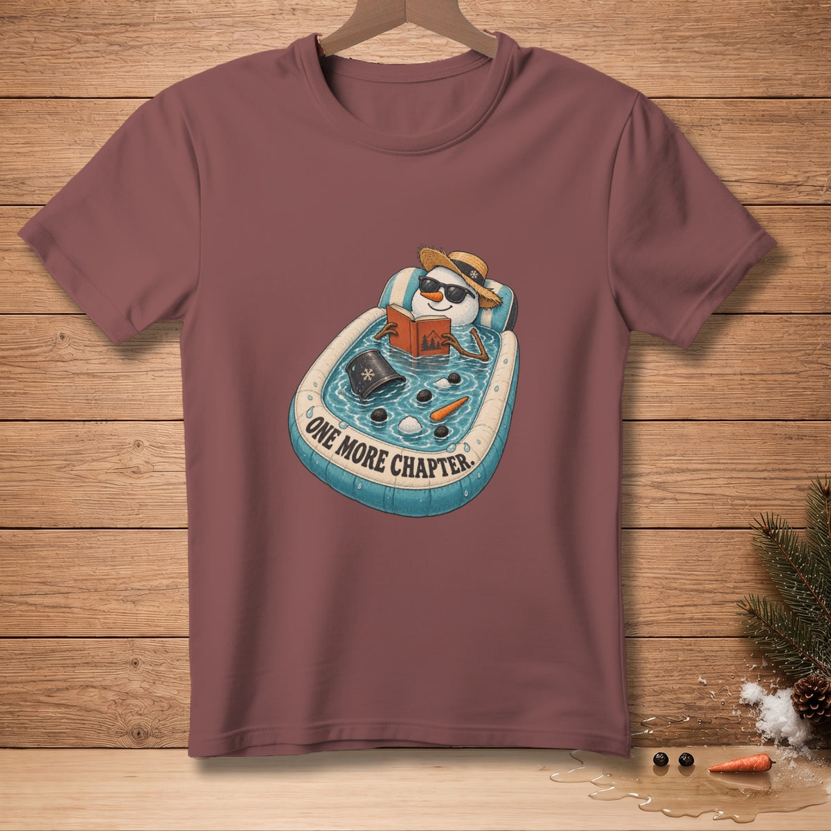 T-shirt graphic of a snowman reading a book in a pool with text "One More Chapter".