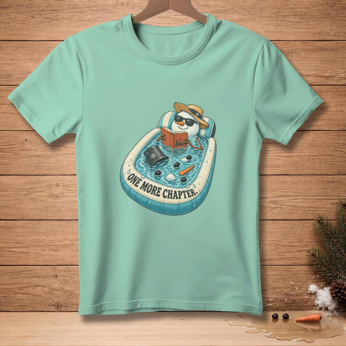 Snowman in pool reading with "ONE MORE CHAPTER" text on a light teal t-shirt.