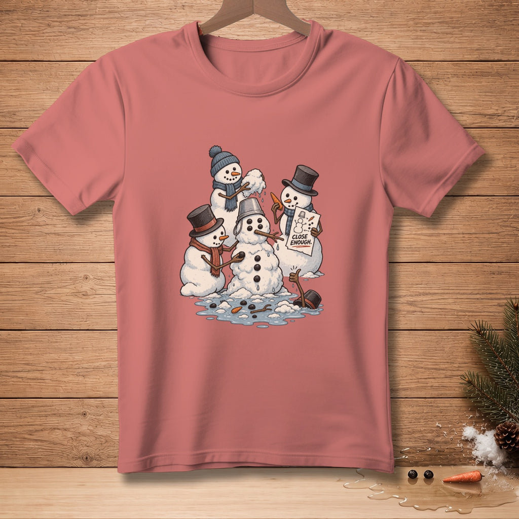 Snowmen building a snowman with "CLOSE ENOUGH" sign on pink t-shirt.
