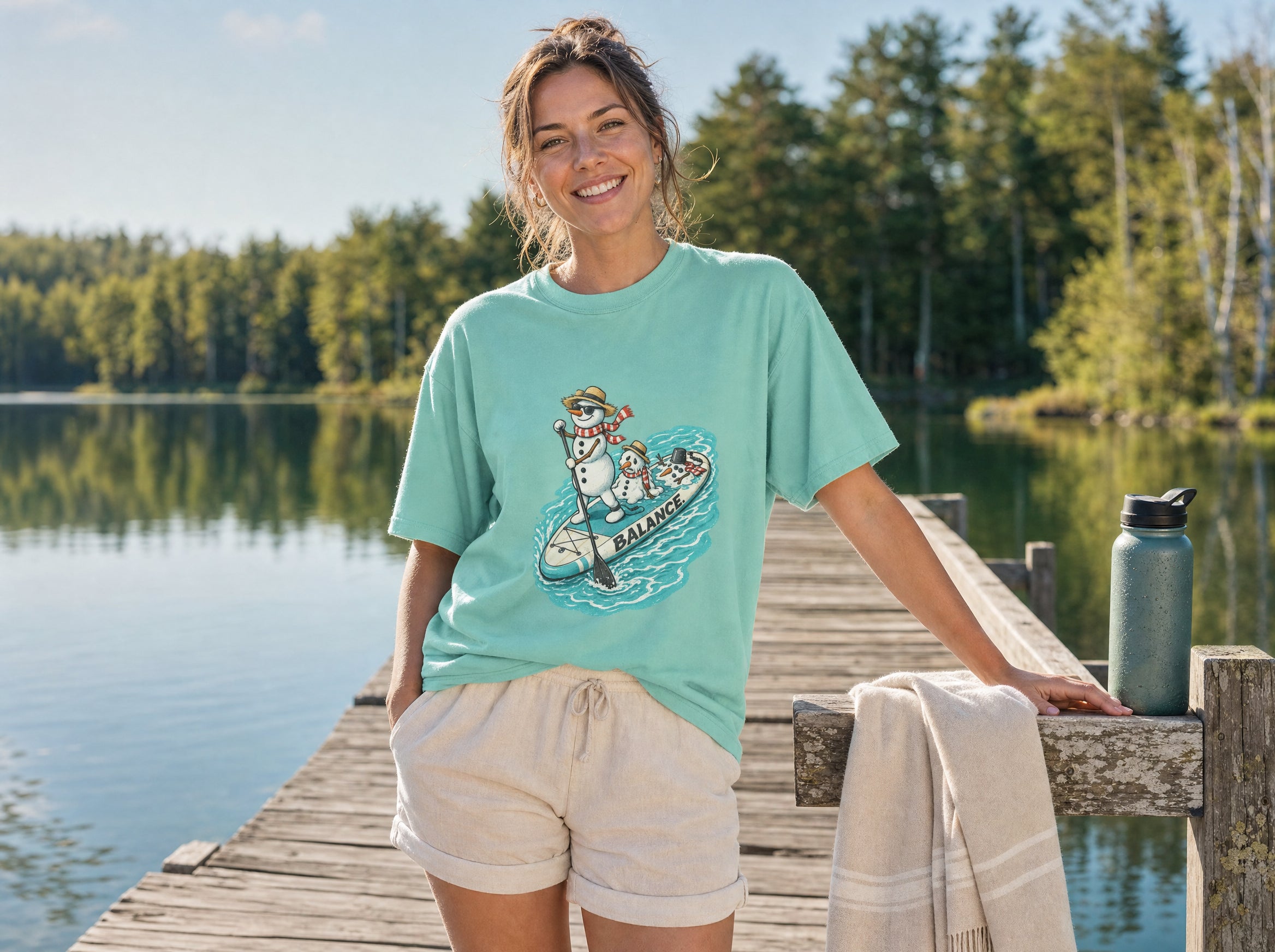A smiling woman on a lake dock wearing the Keep Your Cool Snowman Dayz tee ~ two snowmen on a paddleboard
